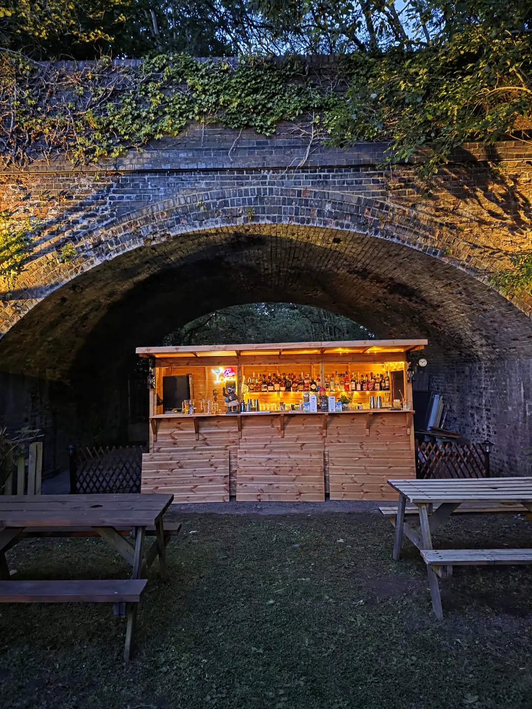 The garden bar glowing under the ivy-covered brick railway arch at dusk, with picnic tables on the grass